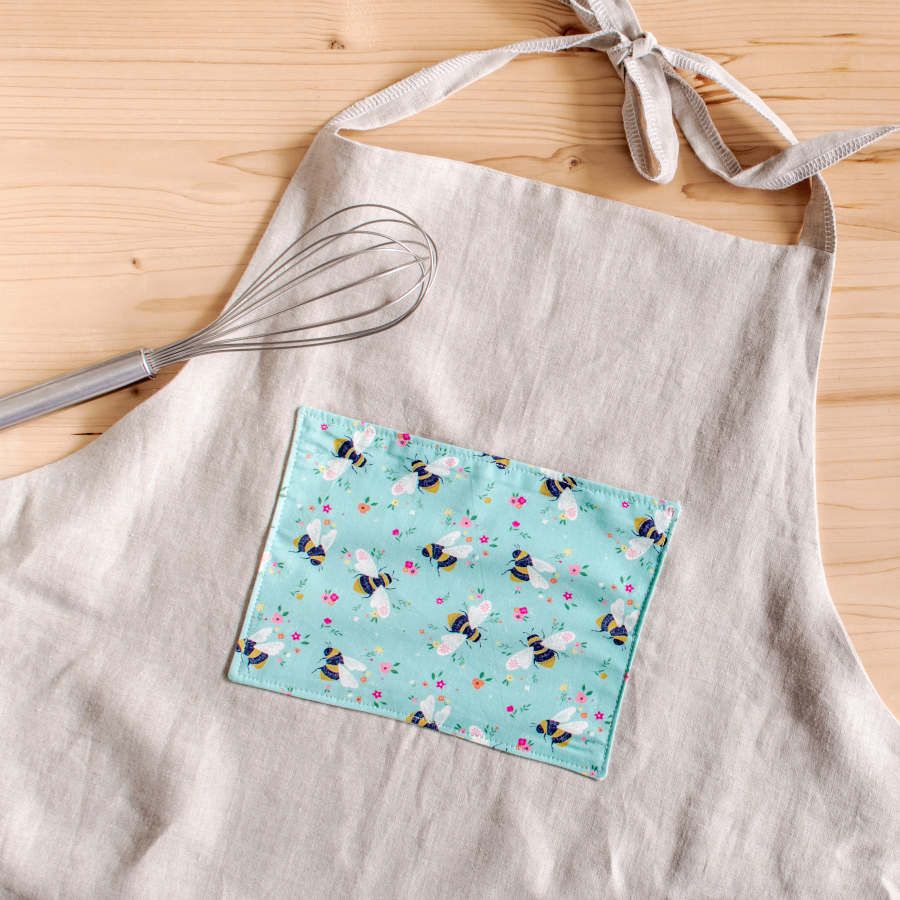 Light grey apron with pocket in the front made of Blue organic cotton poplin with black and yellow bees and colourful flowers